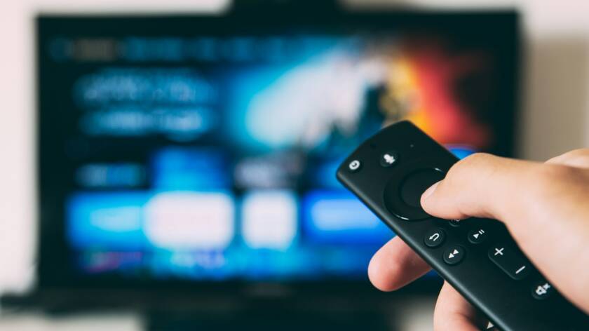 Kuvassa on kaukosäädin ihmisen kädessä. Taustalla näkyy tv-ruutu. Kuvaaja: Glenn Carstens-Peters / Unsplash.